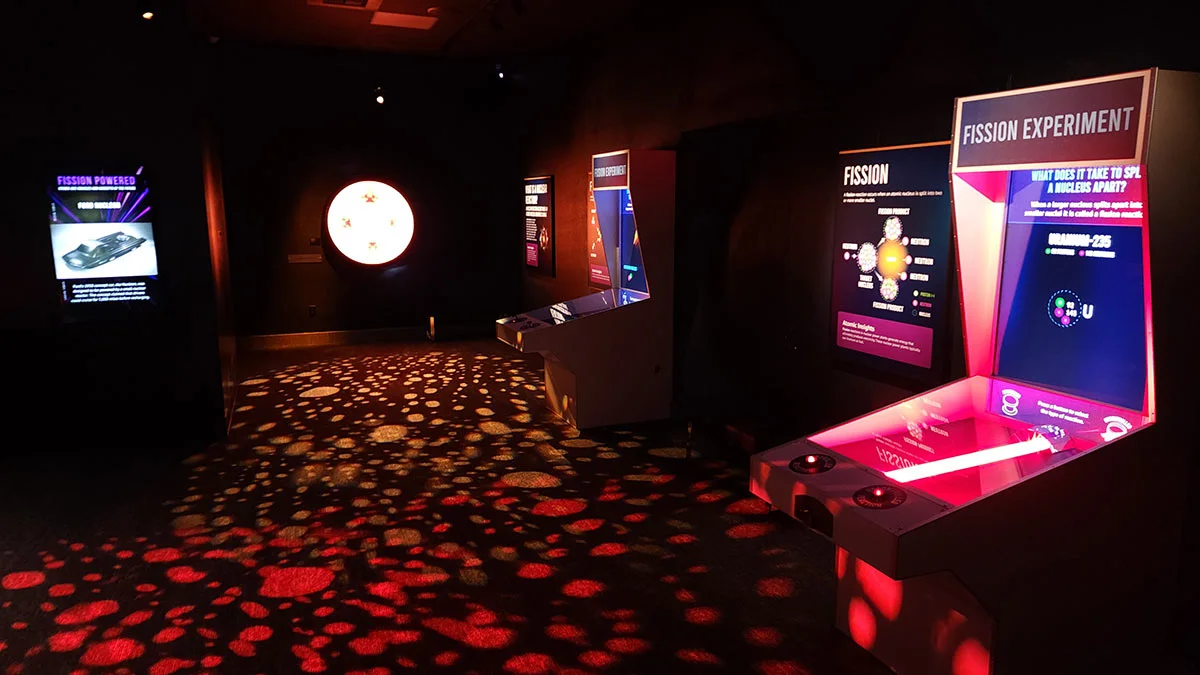 Interactive "Fission Experiment" arcade-style exhibit in a dimly lit museum space.