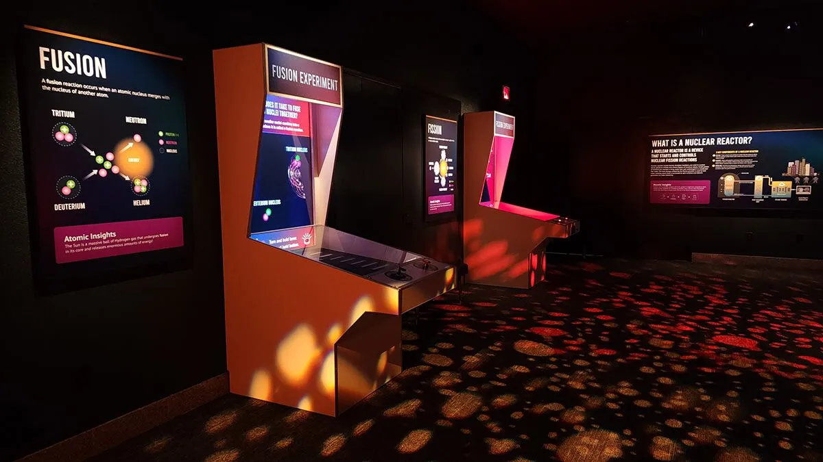 Interactive arcade-style kiosks explaining nuclear fusion and fission reactions in a museum exhibit room.